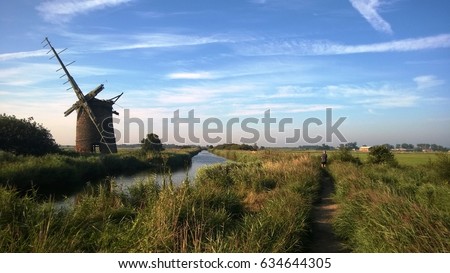 Old windmill by the water on the Norfolk Broads in East Anglia England on a Summer afternoon with blue sky and a man walking in the distance Royalty-Free Stock Photo #634644305