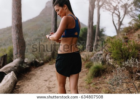 Female runner checking fitness progress on her smart watch. Asian woman using fitness app to monitor workout performance, while walking through mountain trail.