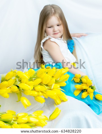 Cute little girl with long blond hair in a long blue skirt sitting on the couch with a large bouquet of bright yellow tulips.