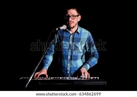Keyboarder in plaid shirt playing and singing on dark background.