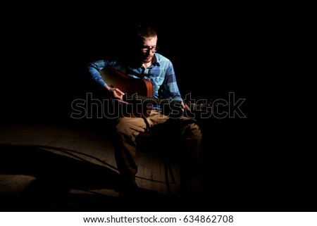 Young musician in plaid shirt playing the guitar and singing on dark background.