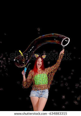 A red headed young girl is surrounded by soap bubbles. She wears 
a pair of flowered ears on her head and plays around with soapy 
water and forms soap bubbles in front of a black background.