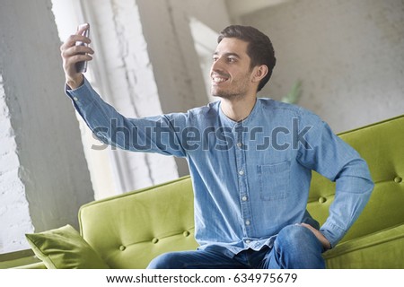 Indoor shot of attractive Caucasian male in nice casual clothes having rest on comfortable sofa, sitting with his cellphone in hand, posing naturally to take selfie picture to post it into web.