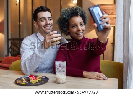 Happy interracial couple dating in cafe in afternoon, dark-skinned woman is stretching her hand holding cellphone to take selfie of her and boyfriend, both are smiling positively and showing content.
