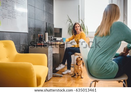 Two woman sitting at the desk and talking in the office and small yellow dog is siting on the floor