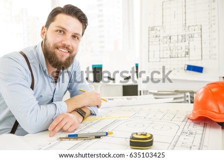 Handsome bearded male architect working on a building plan at his desk smiling to the camera joyfully copyspace professionalism trustworthy qualified experienced engineer constructionist developer Royalty-Free Stock Photo #635103425