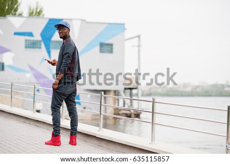 Portrait of stylish african american man on sportswear, cap and glasses with smartphone at hand. Black men model walk street against  lake.