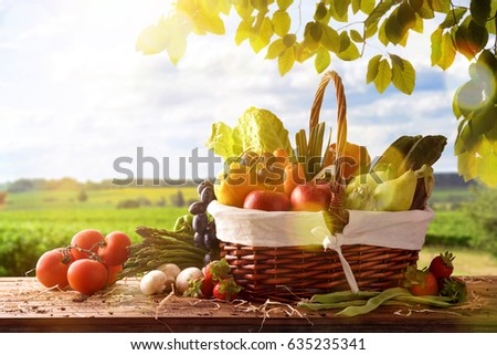 Assortment of fruits and vegetables in a wicker basket on a wooden table on crop landscape background. Horizontal composition. Front view