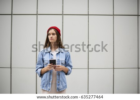 Attractive hipster girl dressed in trendy casual outfit use modern smartphone connected to internet while standing in urban setting.Beautiful stylish dressed female holding cellular against copy space