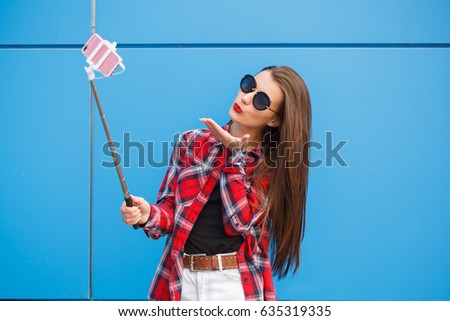Fashion portrait of pretty smiling and woman in sunglasses with smartphone against the colorful blue wall. Make selfie. Kissing