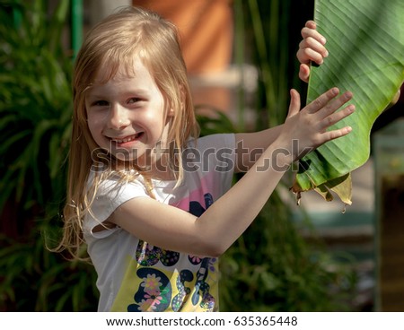 portrait, little girl of seven years, holds large leaf of palm tree, botanical garden, sweet  smiling, long light, wheat hair, happy child, bright day, white T-shirt, sunny, 	
laughing