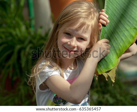 portrait, little girl of seven years, holds large leaf of palm tree, botanical garden, sweet  smiling, long light, wheat hair, happy child, bright day, white T-shirt, head tilted towards plant, sunny