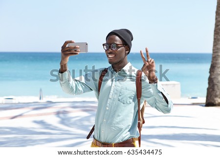 Happy young dark-skinned European male traveler with backpack looking and smiling at camera on his smart phone, showing peace gesture while taking selfie, blue sky and vast ocean in background