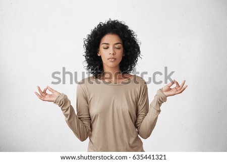 Consideration and praying. Beautiful calm young black female with Afro hairstyle keeping eyes closed while practicing yoga indoors, meditating, holding hands in mudra gesture, thinking about peace