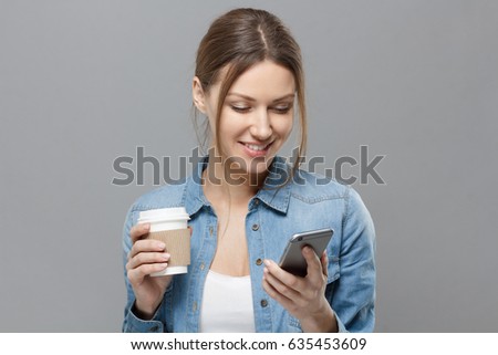 Closeup of young beautiful European lady isolated on gray background holding cardboard coffee cup and smartphone in her hands looking at screen and smiling happily at content she is watching.