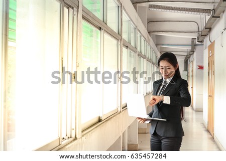 business woman in black suit and tie look at watch on left hand and hold laptop with right hand