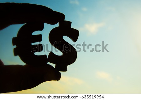 hand hold between two fingers silhouette of share the Dollar and euro sign  icon together - word es . dollar and euro sing against blue and yellow sky background.