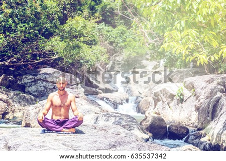 An attractive young man sits in a lotus pose and meditates against a background of stones and a waterfall.