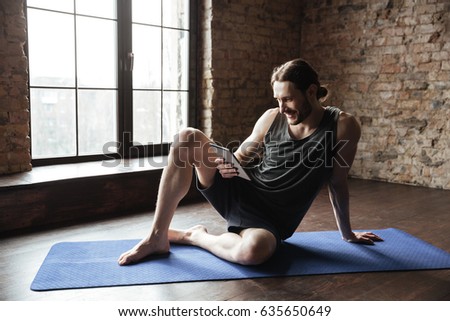 Picture of happy strong sportsman in gym lies on floor chatting by tablet computer. Looking aside.