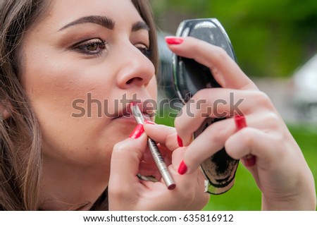 woman corrects make-up and hairstyle in the park. Woman putting, correcting red lipstick lip gloss. 