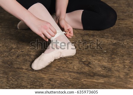 Young ballerina or dancer girl putting on her ballet shoes on the wooden floor. Female dancer ties on her pink ballet slippers with ribbons