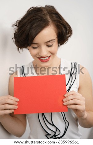 Stylish young woman holding a blank red presentation card with copy space in front of her chest as she looks down at it with a smile