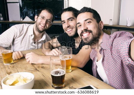 Three young happy friends taking selfie and drinking beer at the pub. Mobile concept