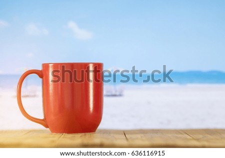red cup of coffee on book at blur white sand beach over blue sky and white lounge chairs with colorful sun umbrella on beach background.