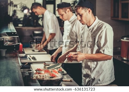 Food concept. Three young chefs in white uniform decorate ready dish in restaurant. They are working on maki rolls. Preparing traditional japanese sushi set in interior of modern professional kitchen