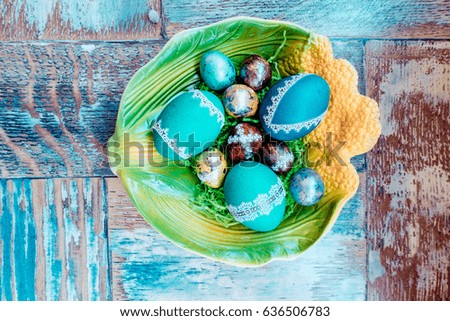 On the old wooden shabby table a dish of metal with painted in a different color Easter eggs chicken and quail and Golden blue green purple feathers, willow