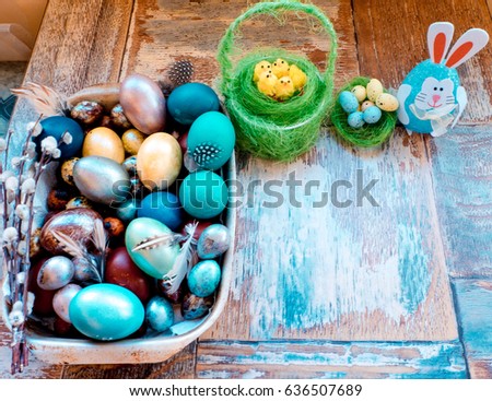 On the old wooden shabby table a dish of metal with painted in a different color Easter eggs chicken and quail and Golden blue green purple feathers, willow and chickens with rabbit