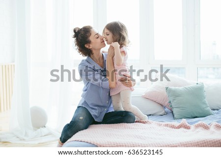 Happy young mother hugging and kissing with her little daughter on the bed in the morning. Enjoying time together at home. Pink and blue pastel colors, selective focus. Love and care concept