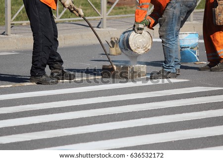 Workers apply a road marking to the pedestrian crossing zebra-stripe crosswalk with white paint and sprinkle the stripes with a reflective powder on the asphalt near the traffic light