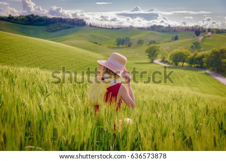 Image of young woman with red ruby dress and hat posing in a field of wheat in Tuscany in spring