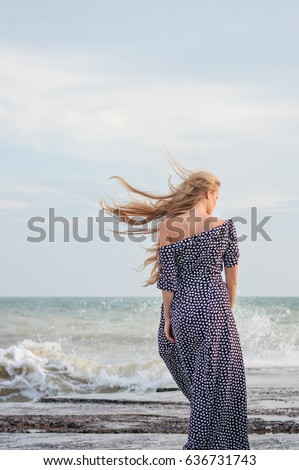 beautiful slender blonde in a long polka-dot dress on a background of azure sea. portrait from the back     Royalty-Free Stock Photo #636731743