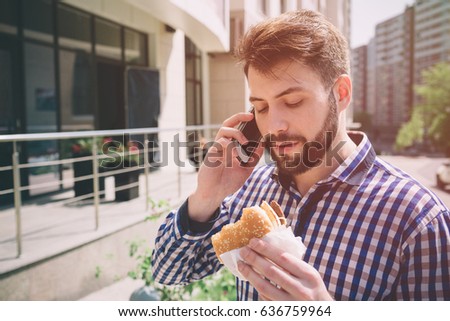 Handsome sporty bearded dark-haired man eating a hamburger and talking on the phone.