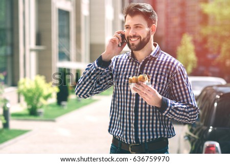 Handsome sporty bearded dark-haired man eating a hamburger and talking on the phone.