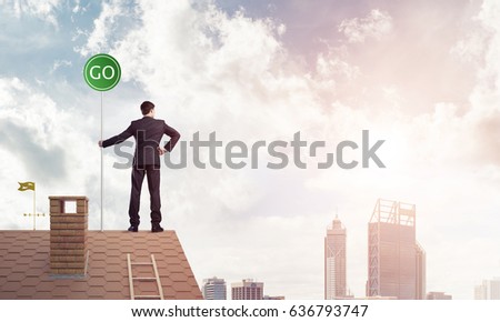Young caucasian businessman standing on house roof and holding go green sign. Mixed media