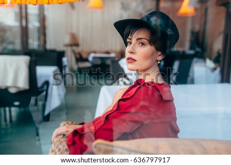 The girl in a black hat and a crimson dress sits in cafe. She thoughtfully looks somewhere in a distance. The girl has red lips, we look at her through a window.