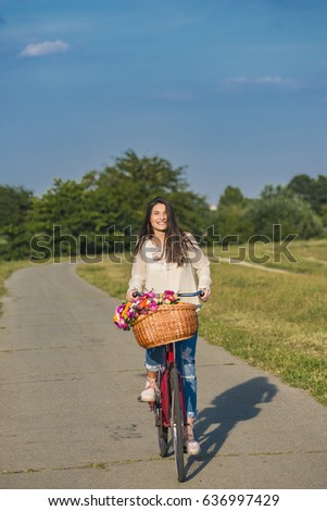 Young smiling woman rides a bicycle with a basket full of flowers in counrtyside