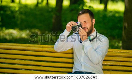 A young guy with a beard sits on a yellow bench in the park and takes pictures.