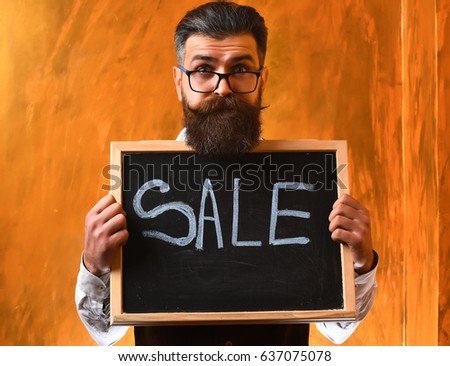 Bearded man, long beard. Brutal caucasian unshaven serious hipster with glasses and moustache holding sale inscription on blackboard on brown studio background