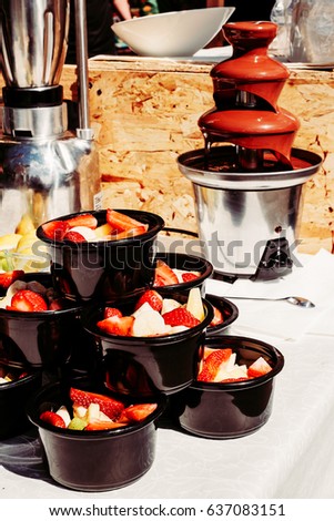 chocolate fountain and slice strawberries banana in black cups on a white table, delicious sweets dessert, street food, outdoor