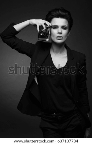 Young serious amazing woman with short dark hair wearing black retro blazer posing in dark studio, holding old photo camera in her hands, in black and white
