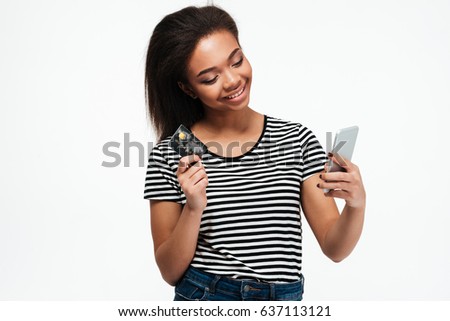 Picture of smiling young african lady standing isolated over white background. Looking aside while using phone and holding debit card.