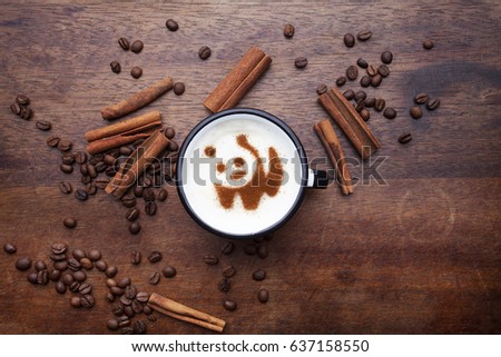 Hand holding a rustic white mug with coffee cream. Food art creative concept image, cute panda bear drawing with cinnamon powder over wooden background.