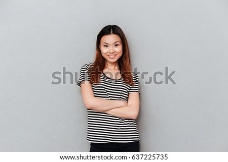 Picture of happy young pretty woman standing over grey wall. Looking at camera with arms crossed.