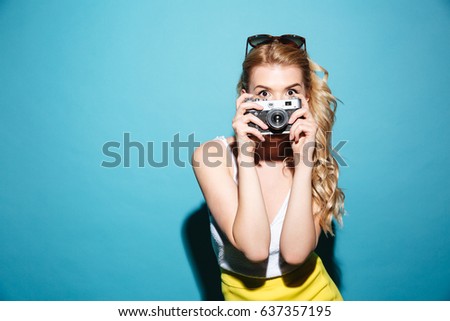 Portrait of a young blonde woman in sunglasses taking picture on retro camera isolated over blue background