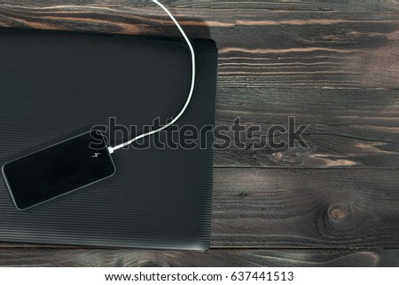 On the wooden table top lies a closed laptop and on top of it is charged a mobile phone with a white charging cable. A table made of brown old wood.