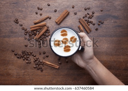 Hand holding a rustic white mug with coffee cream. Food art creative concept image, set of beans drawing with cinnamon powder over wooden background.